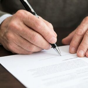 A close-up view shows a senior's hand signing a detailed legal document with a pen.